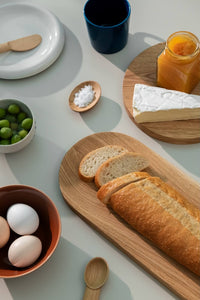 Breakfast table with green olives in a small bowl, navy cup, three eggs in a brown bowl, baguette, brie cheese with some orange jam all placed on the wooden cutting boards of different sizes