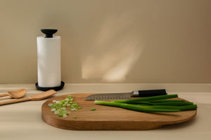 Spring onion and its cut pieces placed on a wooden cutting board, two wooden spatulas and a black paper towel holder in a background