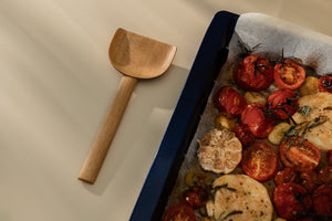 Shovel-like wooden spatula placed alongside baking tray with roasted tomatoes and garlic