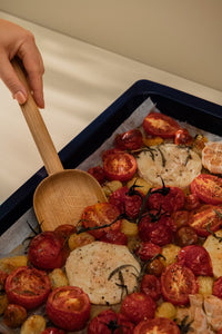Woman scooping roasted tomatoes and garlic from a baking tray with a shovel-like wooden spatula