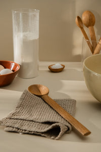 Kitchen counter with a large wooden spoon placed on a grey tea towel alongside large yellow bowl, eggs in a brown bowl, a jar with flour, salt in a small bowl, and some wooden kitchen utensils