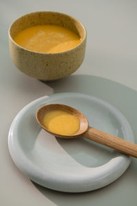 Scoop of pumpkin soup in a wooden spoon placed on a white small plate alongside a bowl full of soup